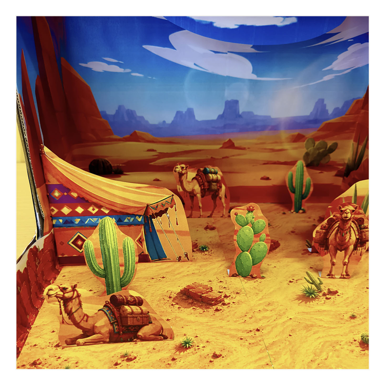 Desert habitat diorama close-up with several camels, green cacti, a colorful desert tent and sandy ground